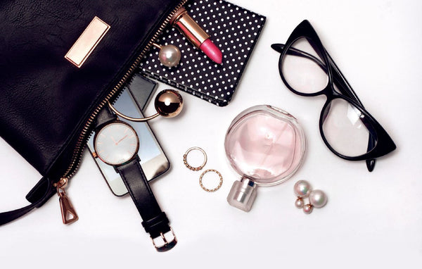Black leather purse with watch, smartphone, lipstick, perfume, glasses, earrings, and a polka dot wallet on a white surface.