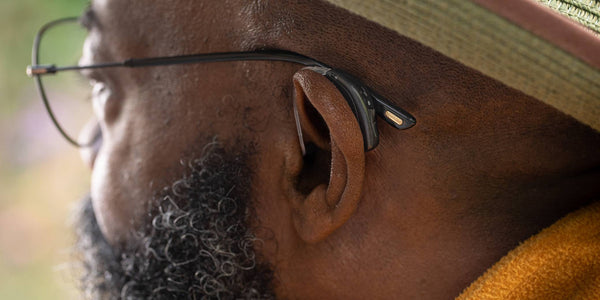 Close-up of a person wearing black-framed glasses and a bone conduction headphone on their ear, with a green hat visible.