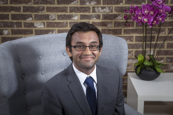 Professional man in a suit sitting in a grey armchair with a brick wall background and purple orchids on a white table nearby.