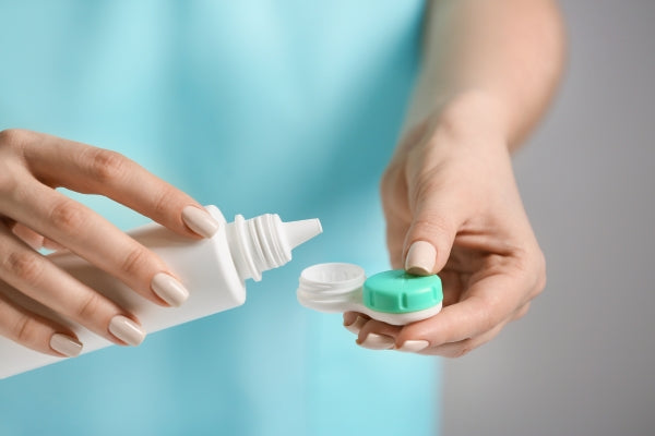 Person pouring contact lens solution into a green and white contact lens case against a blue background.
