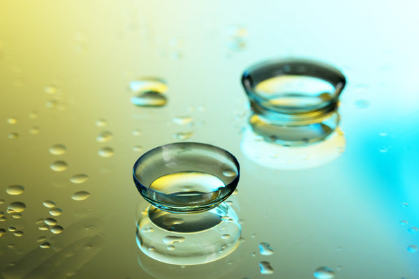 Two contact lenses on a reflective surface with scattered water droplets and a gradient background of yellow and blue tones.