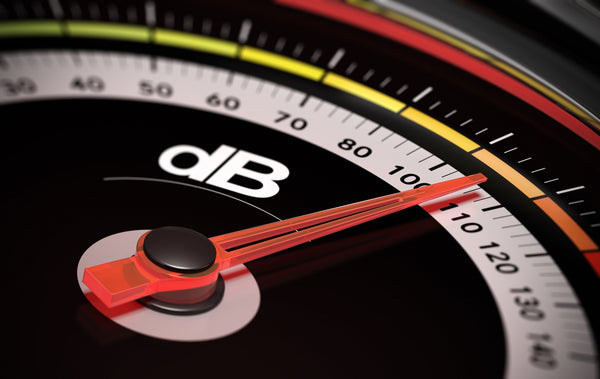 Close-up of a decibel meter needle pointing at over 100 dB, indicating a high sound level with color-coded zones on the gauge.