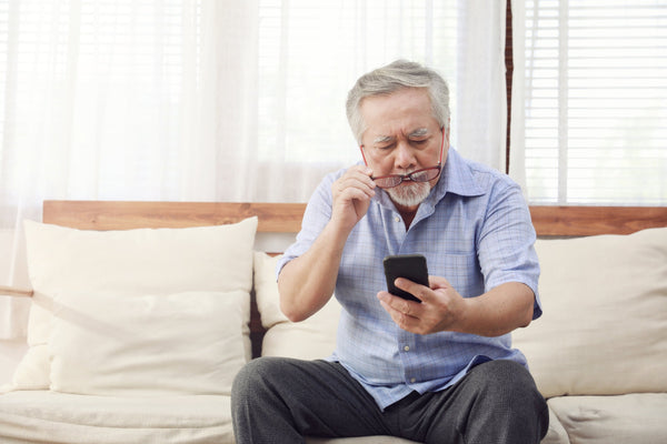 Older man sitting on couch, holding smartphone and adjusting glasses while looking at the screen in a well-lit living room.