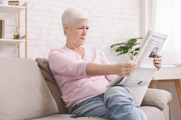Person sitting on a couch reading a newspaper with a focused expression, indoor setting with a plant and shelving in the background.
