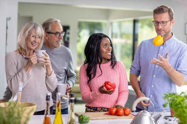 Four adults enjoying time in a bright kitchen preparing vegetables, with a man tossing a yellow bell pepper.