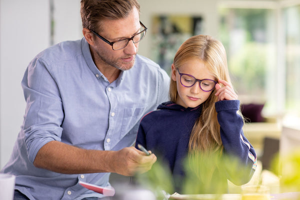 Adult helping child with homework at home, both wearing glasses and focused on the task.