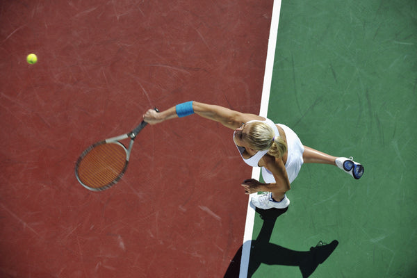 Tennis player in white outfit reaching to hit a tennis ball on an outdoor court with red and green sections.