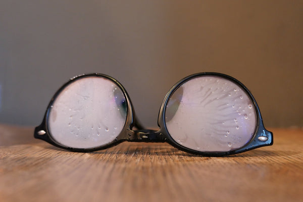 Close-up of black-framed eyeglasses with fogged and water-dotted lenses resting on a wooden surface.
