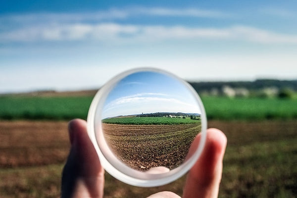 Hand holding a clear glass sphere reflecting a sharp, inverted image of a green agricultural field under a blue sky.