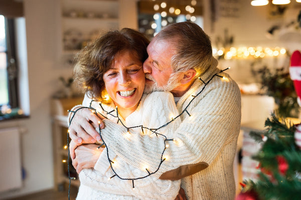 Elderly couple wrapped in holiday string lights, sharing a joyful moment in a cozy, warmly lit home decorated for Christmas.