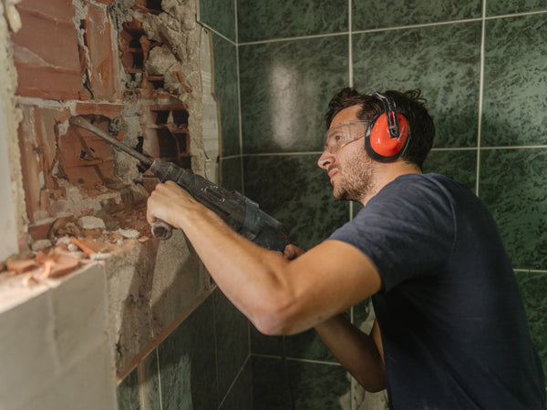 Man using a power drill to break through a tiled wall during home renovation, wearing safety glasses and ear protection.