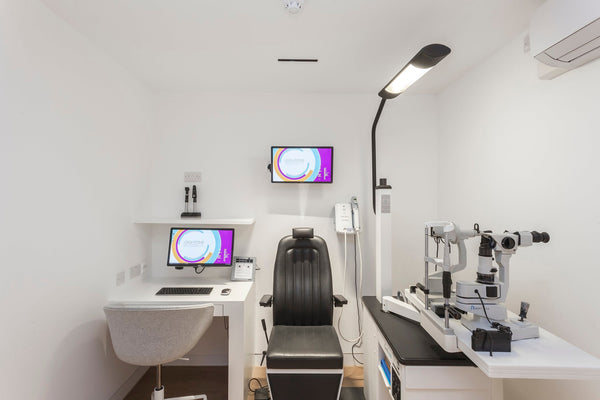 Modern optometry exam room with eye testing equipment, black patient chair, computer monitors, and white walls.