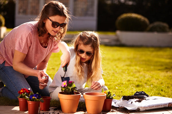 Two people planting flowers in pots outdoors on a sunny day, surrounded by gardening supplies and soil.