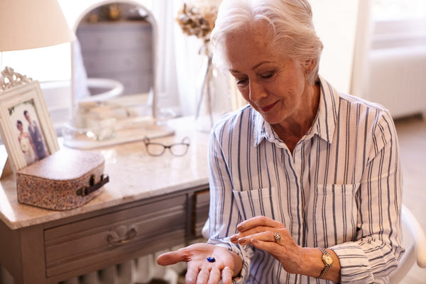 Older person holding and preparing to take a blue pill while sitting at a desk with glasses and a photo frame in the background.