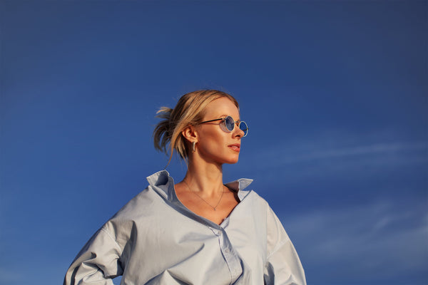 Stylish woman wearing round sunglasses and a light blue oversized shirt, posing confidently under a clear blue sky — summer fashion and eyewear inspiration.