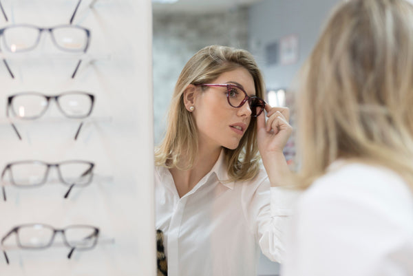 Woman trying on eyeglasses in front of a mirror at an optical store, with multiple glasses displayed on a nearby rack.