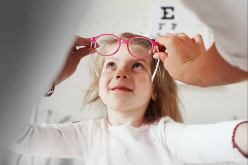 Child wearing pink glasses with an adult's hand adjusting them, against a neutral background.