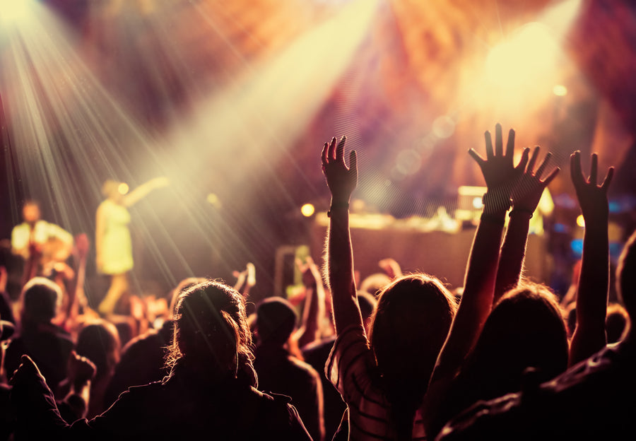 Crowd with raised hands enjoying a live concert with bright stage lights and a performer in the background.