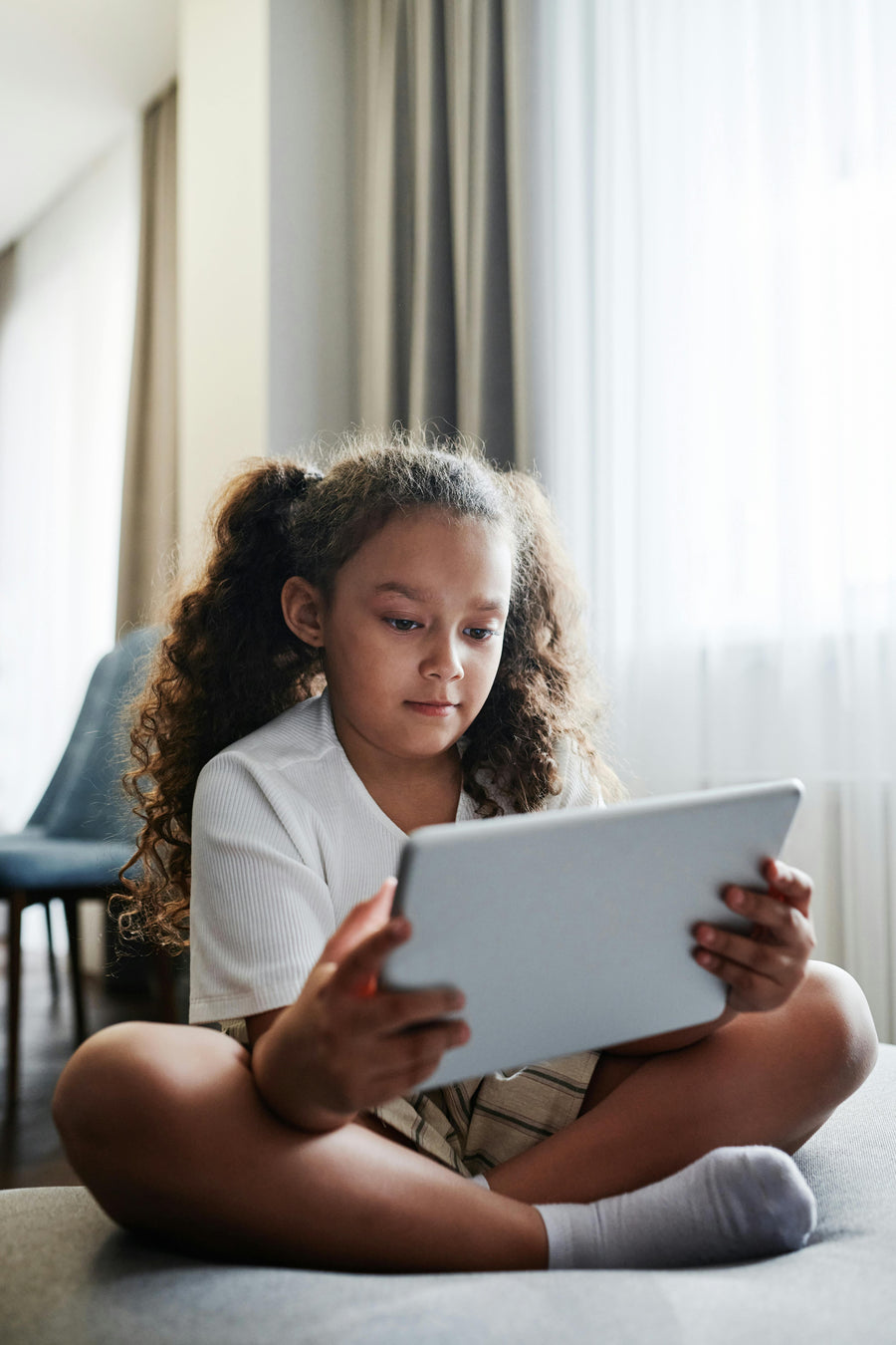Young girl sitting on a couch using a tablet in a bright room.