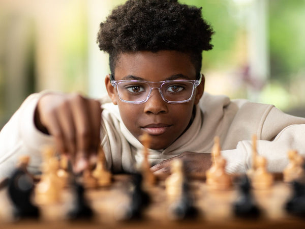 Young person wearing glasses focused on moving a chess piece on a wooden chess board indoors with natural background light.