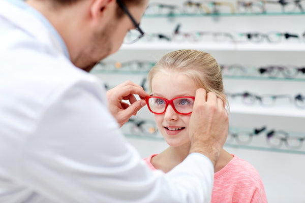 Optician helping a young girl try on red eyeglass frames in an optical store.