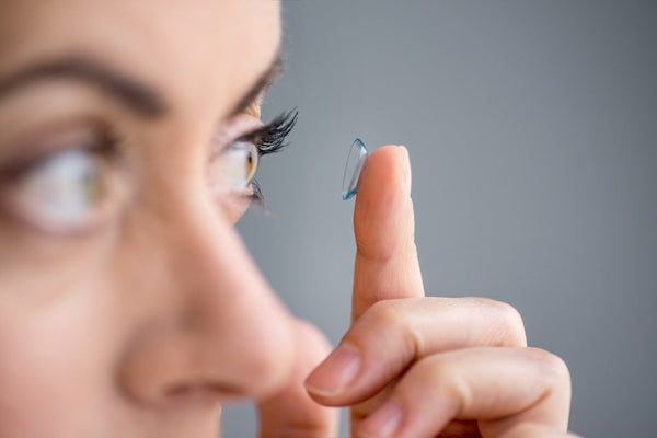 Close-up of a person holding a contact lens on their fingertip, about to place it in their eye against a gray background.