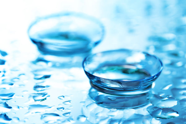 Two clear contact lenses resting on a wet, reflective blue surface with water droplets scattered around.