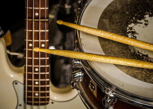 Close-up of drumsticks resting on a worn drumhead beside the neck of an electric bass guitar in a music setup.
