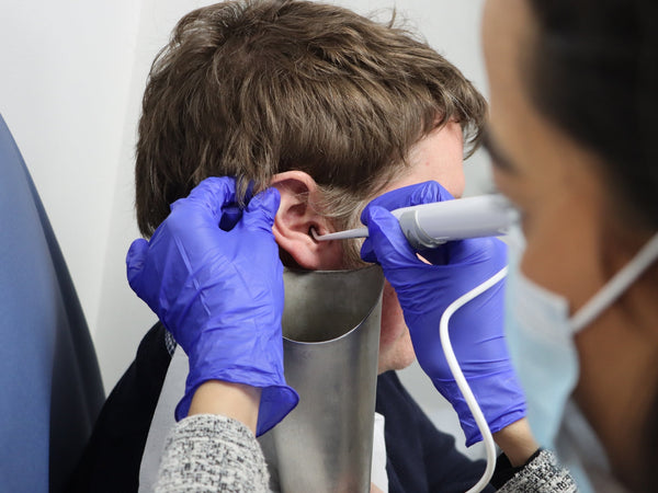 Medical professional wearing gloves and a mask using an otoscope to examine a patient's ear in a clinical setting.