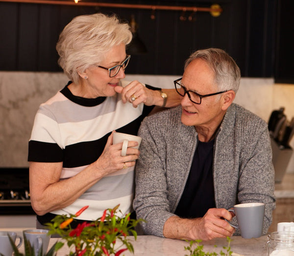 Older couple enjoying coffee together in a modern kitchen, surrounded by plants and warm lighting.