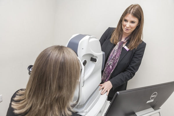 Optometrist assisting a patient with eye examination using a specialized diagnostic machine in a clinical setting.