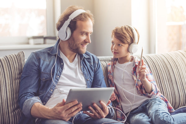Man and child sitting on a couch, wearing headphones and sharing audio from a tablet and smartphone with a connected cable.