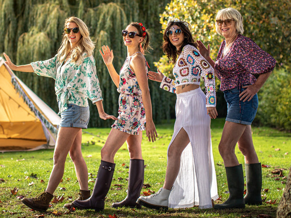 Four people in colorful outfits and sunglasses walking in a park with green grass and trees, next to a yellow tent.