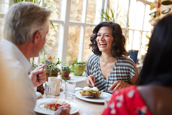 Three people enjoying a meal together and laughing at a bright, plant-filled dining space.
