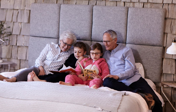 Grandparents and grandchildren sitting on bed together, reading storybooks in a cozy bedroom setting.