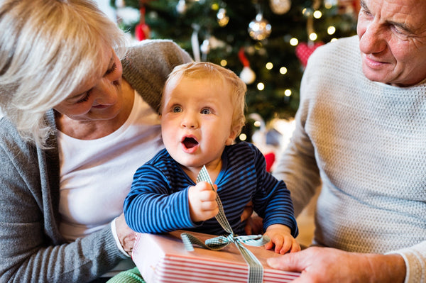 Child sitting with elderly adults near a Christmas tree, holding and opening a wrapped gift with a ribbon bow.