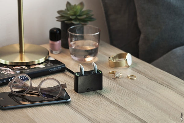 Desk setup with wireless hearing aids in charging case, eyeglasses, smartphone, watch, rings, nail polish, a book, glass of water, and a small plant.