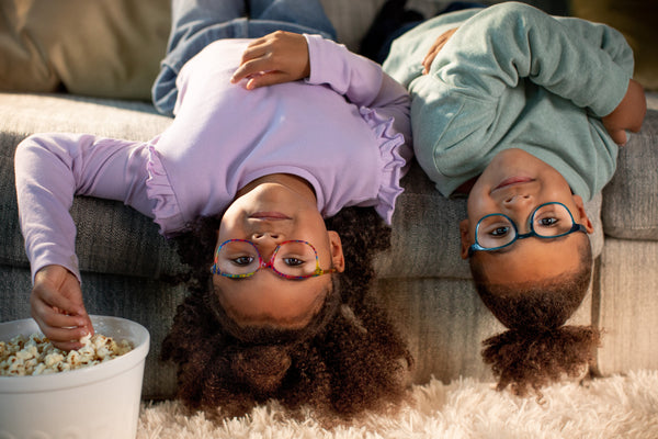 Two children wearing glasses lying upside down on a couch with a bowl of popcorn nearby.