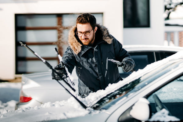 Person in winter jacket removing snow from a car windshield with a snow brush and ice scraper on a snowy day.