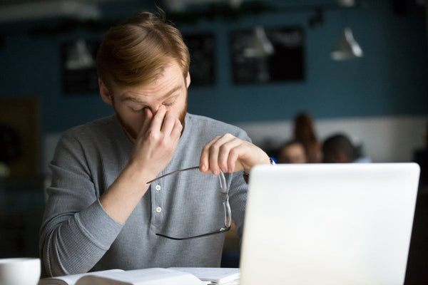 Person removing glasses and rubbing eyes while sitting at a desk with a laptop and open book in a dimly lit room.