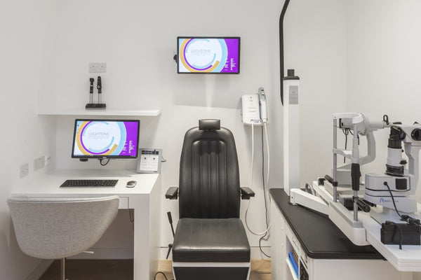 Modern optometry exam room with black patient chair, diagnostic equipment, wall-mounted screens, and white desk with computer.