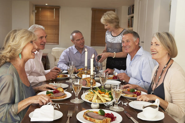 Group of people enjoying a festive meal with meat, vegetables, wine, and candles around a dining table in a home setting.