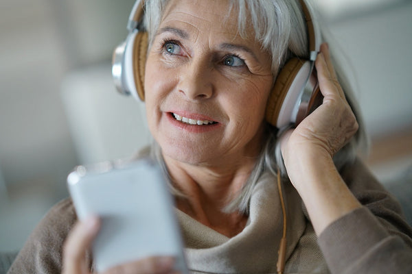Older person wearing headphones and holding a smartphone, enjoying music or a podcast indoors with a relaxed expression.