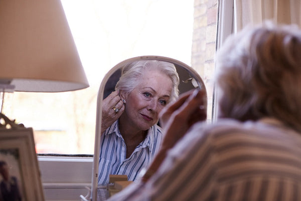 Older woman adjusting earrings while looking into a mirror on a dressing table near a window with natural light.