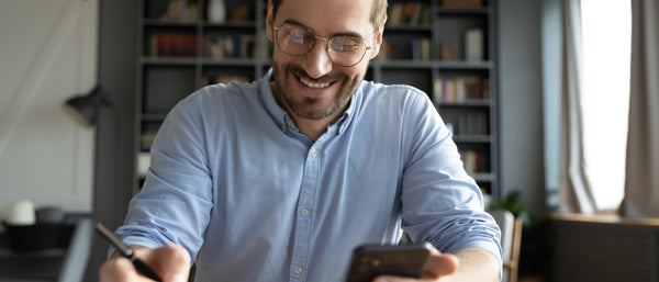 Man in glasses smiling while using a smartphone and writing with a pen in a home office with bookshelves in the background.
