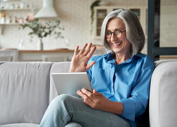 Older adult sitting on a couch and smiling while waving at a tablet during a video call in a cozy living room.