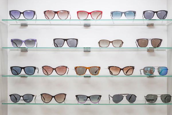 Display of various stylish sunglasses in different colors and designs on glass shelves against a white wall.