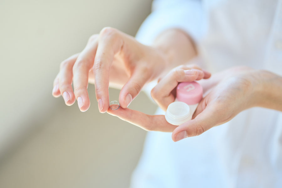 Person holding a contact lens on a fingertip and a small contact lens case with pink and white lids.
