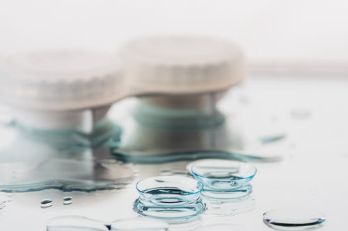 Contact lenses, a white storage case, and a dropper bottle dispensing liquid on a white surface.