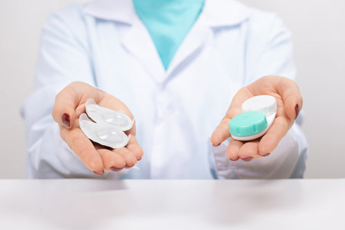 Hands holding contact lenses in blister packs and a contact lens case, with a person wearing a lab coat in the background.